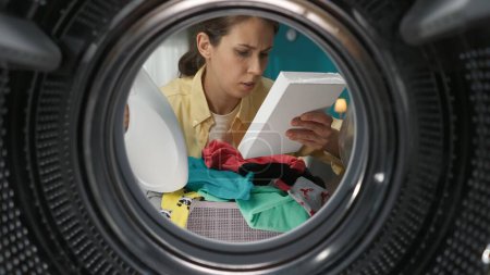 Photo for A portrait of a young adult woman in casual clothing reading instructions on the box of washing powder. View from inside the washing machine. - Royalty Free Image