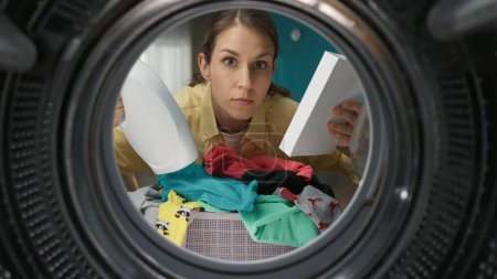 A portrait of a young adult woman in casual holding the box of washing powder and fabric softener. View from inside the washing machine.