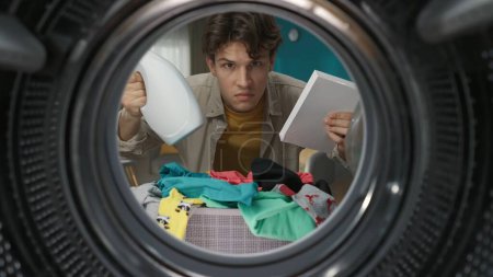 Téléchargez les photos : Portrait d'un jeune homme adulte vêtu de vêtements décontractés tenant la boîte de poudre à laver et d'adoucissant à l'air confus. Vue de l'intérieur de la machine à laver. - en image libre de droit