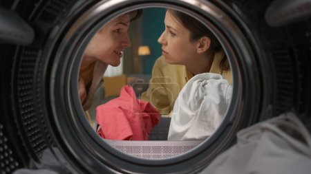 Téléchargez les photos : Portrait d'un jeune homme et d'une jeune femme se regardant et tenant un vêtement dans leurs mains. Couple faisant la lessive ensemble. Vue de l'intérieur de la machine à laver. - en image libre de droit