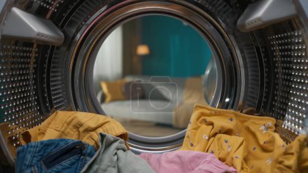 Photo for A closeup shot of Washing machine full of colorful clothes inside the drum with opened door. Laundry daily routine concept. View from inside the washing machine into an empty living room. - Royalty Free Image