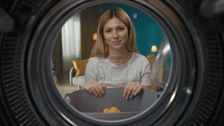 Photo for Adult woman in casual clothing with laundry basket full of colorful clothes. She looks happy and smiles to the camera. View from inside the washing machine. - Royalty Free Image