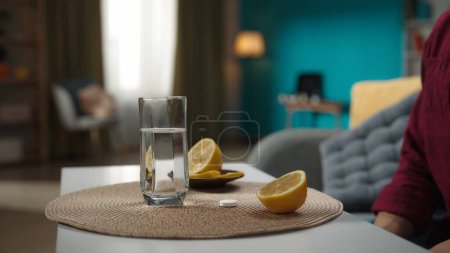 Photo for A close up shot of the woman sitting on the couch, glass of water standing on the table, lemon and effervescent pill lying next to it. Home medicine concept. Healthcare advertisement. - Royalty Free Image