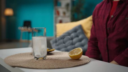 Photo for A close up shot of the woman sitting on the couch, waiting for effervescent pill to dissolve in the glass of water,lemon is lying next to it. Home medicine concept. Healthcare advertisement. - Royalty Free Image