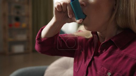 Photo for Close up shot of an adult blonde woman sitting on the sofa, having an asthma attack. Woman using an inhaler. Asthma treatment. Healthcare advertisement. - Royalty Free Image