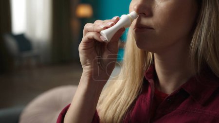 Photo for Portrait of a blonde woman model sitting on the sofa. Taking a nasal spray and spraying it inside her nose. Rhinitis treatment. Home medicine concept. - Royalty Free Image