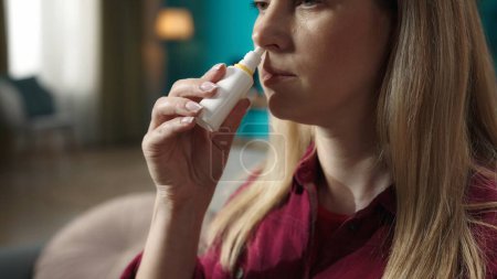 Photo for Close up shot of a blonde woman model sitting on the sofa. Taking a nasal spray and spraying it into her nose. Rhinitis treatment. Home medicine concept. - Royalty Free Image