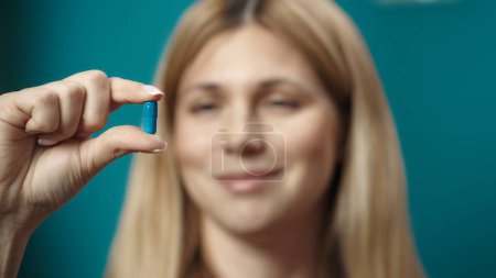 Photo for Close up shot of female blonde model holding a blue capsule pill in her hand, looking at it and smiling. Home medicine concept. Healthcare advertisement. - Royalty Free Image