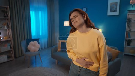 A woman holds her hand on her stomach, suffering from pain, standing in the living room. A woman suffers from abdominal pain, menstrual pain, indigestion, diarrhea. Home medicine concept
