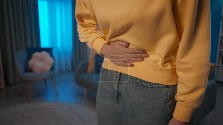Photo for A woman is in pain while standing in the living room. A womans hand is holding on to her stomach close up. A woman suffers from abdominal pain, menstrual pain, indigestion, diarrhea. Home medicine - Royalty Free Image