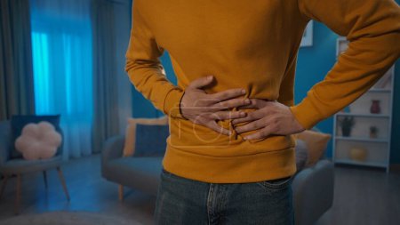Photo for A man is experiencing pain in his stomach while standing in the living room. Hands of a man holding his stomach close up. A man suffers from abdominal pain, indigestion, diarrhea. Home medicine - Royalty Free Image