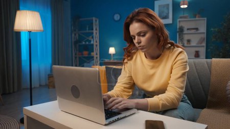 Téléchargez les photos : Une femme travaille sur un ordinateur portable alors qu'elle est assise sur le canapé dans le salon le soir. Une femme vérifie email, types, joue en ligne - en image libre de droit