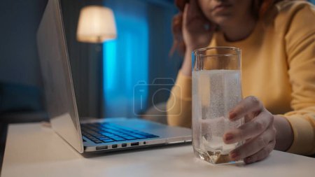 Photo for The womans hand reaches for a glass of water, in which an effervescent tablet dissolves close up. A woman suffers from a headache while working on a laptop. Cure for a cold, pain or hangover. Home - Royalty Free Image