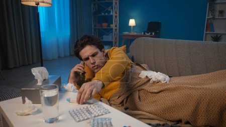 Foto de Un hombre enfermo yace en el sofá y llama a su teléfono inteligente, mira el termómetro. Un hombre llama a una ambulancia o a un médico. Concepto de medicina casera - Imagen libre de derechos