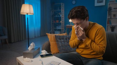 Photo for A man with cold symptoms is sitting on a sofa in the living room. A man blows his nose into a paper handkerchief. Colds, nasal congestion, allergies. Home medicine concept - Royalty Free Image