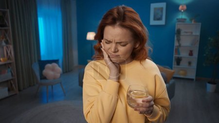 Eine junge Frau steht im Wohnzimmer und hält ihre Wange in Großaufnahme. Eine Frau leidet unter Zahnschmerzen, nachdem sie kaltes Wasser mit Eis und Zitrone getrunken hat. Zahnempfindlichkeit, Zahnschmerzen. Hausmedizin