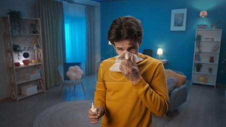 Photo for A man with symptoms of a cold stands in the living room close up. A man blows his nose into a paper handkerchief and holds a nasal spray. Home medicine concept - Royalty Free Image