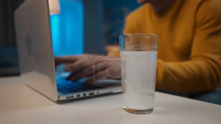 Photo for An effervescent tablet dissolves in a glass of water on a table close up. In the background, a mans hands are typing on a laptop. Cure for a cold, pain or hangover. Home medicine concept - Royalty Free Image