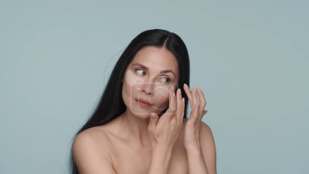 Téléchargez les photos : Femme brune faisant des procédures de soins de la peau faciale. Une femme touche les taches hyaluroniques sous ses yeux, les lissant sur un fond bleu de près. Le concept de beauté, rajeunissement, cosmétologie - en image libre de droit