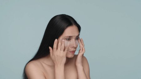 Téléchargez les photos : Femme brune faisant des procédures de soins de la peau faciale. Une femme touche les taches hyaluroniques sous ses yeux, les lissant sur un fond bleu de près. Le concept de beauté, rajeunissement, cosmétologie - en image libre de droit