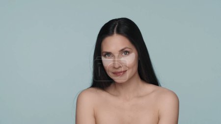 Téléchargez les photos : Femme brune faisant des procédures de soins de la peau faciale. Portrait d'une séminude avec des patchs hyaluroniques sous les yeux en studio sur fond bleu. Le concept de beauté, rajeunissement - en image libre de droit