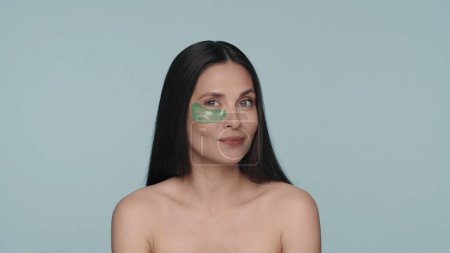Téléchargez les photos : Femme brune faisant des procédures de soins de la peau faciale. Portrait d'une femme avec un patch vert hydrogel sous l'œil en studio sur un fond bleu en gros plan. Le concept de beauté, rajeunissement - en image libre de droit