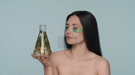 Photo for A woman with green hydrogel patches under her eyes holds a flask with a plant. Seminude woman in the studio on a blue background. Cosmetic products based on natural plant ingredients. The concept of - Royalty Free Image