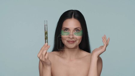 Téléchargez les photos : Une femme avec des taches d'hydrogel vert sous les yeux tient une éprouvette avec une plante. Séminude femme dans le studio sur un fond bleu. Produits cosmétiques à base d'ingrédients végétaux naturels. Le concept - en image libre de droit