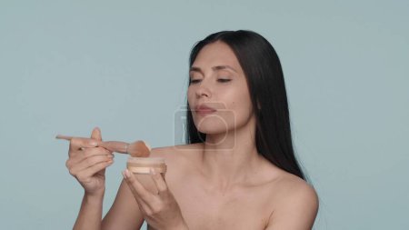 Photo for A woman dips a makeup brush into a powder jar. A woman with cosmetics to improve the contour of the face on a blue background. Contouring, applying powder, blush. The concept of beauty, cosmetology - Royalty Free Image