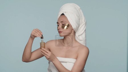 Foto de Una mujer envuelta en toallas de baño después de una ducha, con manchas doradas bajo sus ojos, realiza tratamientos de belleza después de una ducha. Una mujer sostiene una botella de aceite esencial y una pipeta en sus manos. El - Imagen libre de derechos