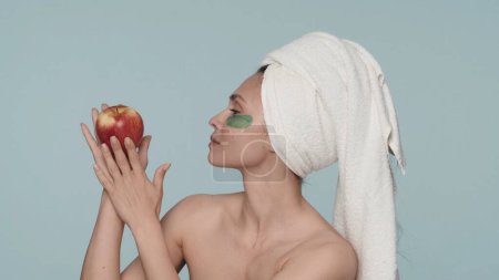 Téléchargez les photos : Femme séminude avec une serviette sur la tête et des taches d'hydrogel vert sous ses yeux sur un fond bleu. Une femme tient une pomme rouge mûre dans sa main. Le concept de beauté, cosmétologie, soin de la peau - en image libre de droit