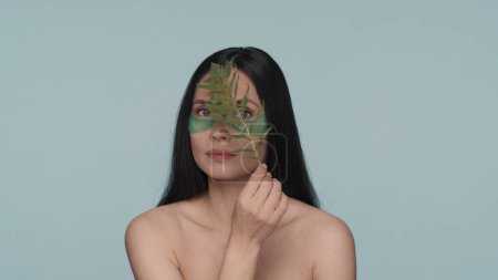 Téléchargez les photos : Une femme séminude avec des taches hyaluroniques sur les yeux court un brin de plante sur son visage. Femme en studio sur fond bleu de près. Cosmétiques naturels à base de plantes. Le concept de beauté - en image libre de droit