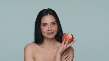 Téléchargez les photos : Une femme avec des taches rouges hydrogel sous les yeux tenant une pomme mûre dans ses mains. Seminude brunette femme dans le studio sur un fond bleu gros plan. Le concept de beauté, rajeunissement - en image libre de droit