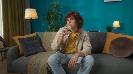 Photo for Closeup shot portrait of a young male model in casual clothing sitting on the sofa in the living room and drinking a glass of water. Leisure time at home. - Royalty Free Image