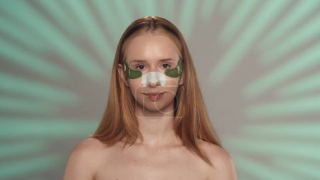 Téléchargez les photos : Une femme avec des taches sous les yeux et un plâtre de points noirs dans le studio sur un fond vert avec des reflets. Le concept de beauté, de cosmétologie, de soins - en image libre de droit