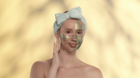 Téléchargez les photos : Une femme applique un masque cosmétique vert sur son visage. Seminude femme avec un groupe de cheveux dans le studio sur un fond jaune avec des faits saillants. Soins de beauté et traitement - en image libre de droit