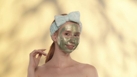 Téléchargez les photos : Une jeune femme avec un bandeau sur la tête effectue des soins cosmétiques de la peau. Portrait d'une séminude avec un masque vert sur le visage en studio sur fond jaune avec des reflets - en image libre de droit