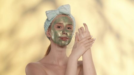 Téléchargez les photos : Une femme avec un masque cosmétique vert sur le visage et un bandeau en studio sur fond jaune avec des reflets. Femme appliquant de la crème pour les mains tout en prenant soin de sa peau. Le concept de beauté - en image libre de droit