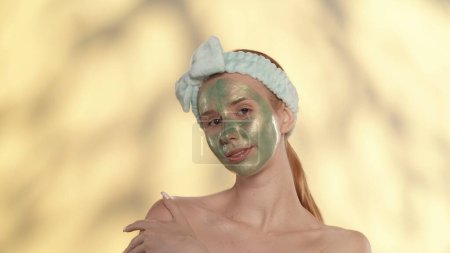 Téléchargez les photos : Une jeune femme avec un bandeau sur la tête effectue des soins cosmétiques de la peau. Portrait d'une séminude avec un masque vert sur le visage en studio sur fond jaune avec des reflets - en image libre de droit