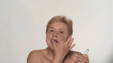 Téléchargez les photos : Portrait d'une femme âgée. Gros plan d'une mannequin femme d'âge moyen appliquant un produit cosmétique sur sa peau, souriant à la caméra. Beauté concept de publicité de soins de la peau. - en image libre de droit