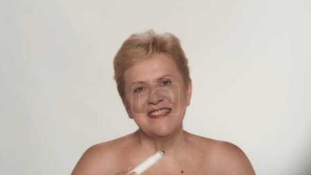 Téléchargez les photos : Portrait d'une femme âgée. Gros plan d'une mannequin femme d'âge moyen qui applique un produit cosmétique sur sa peau, en regardant la caméra. Publicité beauté anti vieillissement soin de la peau. - en image libre de droit