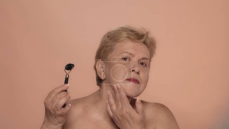 Téléchargez les photos : Portrait d'une femme âgée. Gros plan d'un mannequin féminin d'âge moyen faisant un massage avec un rouleau et regardant la caméra. Publicité beauté anti-âge. - en image libre de droit