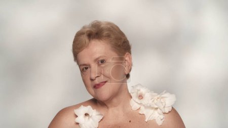Photo for Portrait of a happy senior woman. Close up shot of a middle aged female model posing, smiling at the camera with a white gladiolus flowers on her shoulders. Beauty concept. - Royalty Free Image