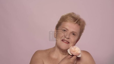 Photo for Portrait of a beautiful senior woman. Close up shot of a middle aged female model holding a rose flower and smiling naturally at the camera. Beauty skincare advertisement concept. - Royalty Free Image