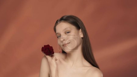 Téléchargez les photos : Portrait d'une jeune femme tenant un bouton de rose rouge dans sa main en gros plan. Une séminude dans le studio sur fond rouge. Le concept de beauté, de cosmétologie, de soins - en image libre de droit