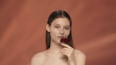 Téléchargez les photos : Portrait d'une jeune femme tenant un bouton de rose rouge dans sa main en gros plan. Une séminude dans le studio sur fond rouge. Le concept de beauté, de cosmétologie, de soins - en image libre de droit