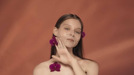 Téléchargez les photos : Une jeune femme avec des fleurs d'orchidée dans ses cheveux, près de son visage et sur sa poitrine. Portrait d'une séminude avec des fleurs d'orchidée en studio sur fond rouge. Le concept de beauté, de cosmétologie - en image libre de droit