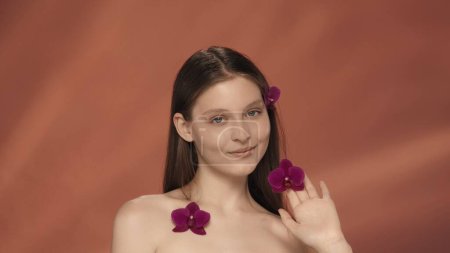 Foto de Una joven con flores de orquídea en el pelo, cerca de la cara y en el pecho. Retrato de una mujer seminude con flores de orquídea en el estudio sobre un fondo rojo. El concepto de belleza, cosmetología - Imagen libre de derechos