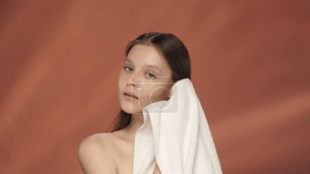 Téléchargez les photos : Tellement doux. Une jeune femme essuie son visage et ses cheveux avec une serviette éponge, appréciant le toucher doux. Séminude femme dans le studio sur un fond rouge. Le concept de beauté, soin de la peau - en image libre de droit