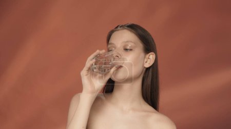 Téléchargez les photos : Une jeune femme boit de l'eau propre et claire dans un verre. Séminude femme dans le studio sur un fond rouge. Maintenir l'équilibre hydrique dans le corps, un mode de vie sain. Le concept de beauté, de cosmétologie - en image libre de droit